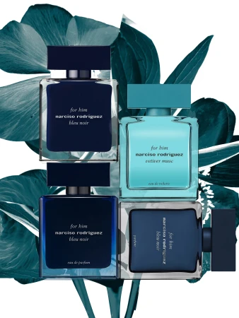 Still life of Narciso Rodriguez 'for him' Bleu Noir and Vetiver Musc fragrances with blue and green accent leaves on a white backdrop.