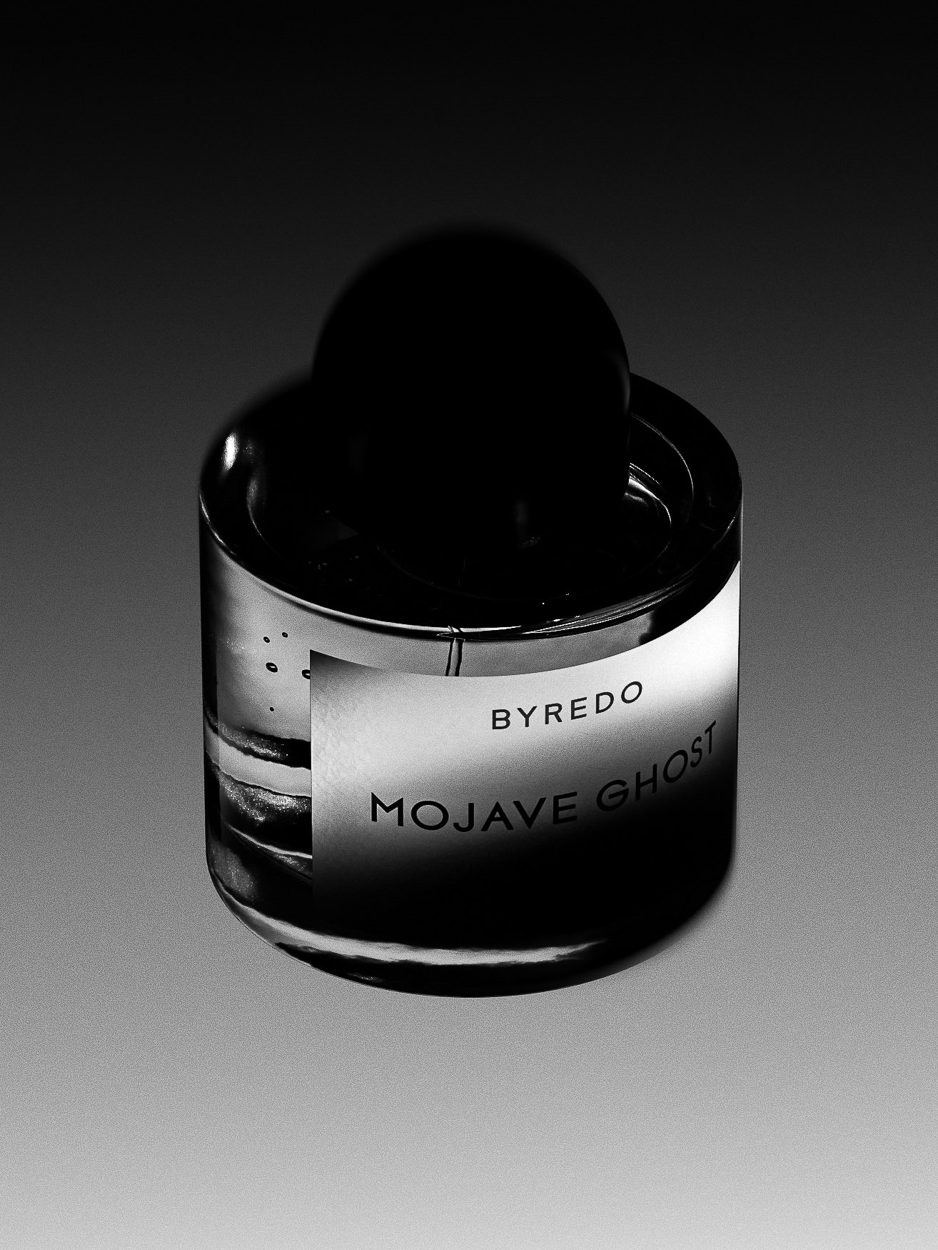 Studio shot of a perfume bottle with a cylindrical body, a domed black cap, and a white label with 'BYREDO MOJAVE GHOST' printed on it.