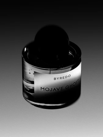 Studio shot of a perfume bottle with a cylindrical body, a domed black cap, and a white label with 'BYREDO MOJAVE GHOST' printed on it.
