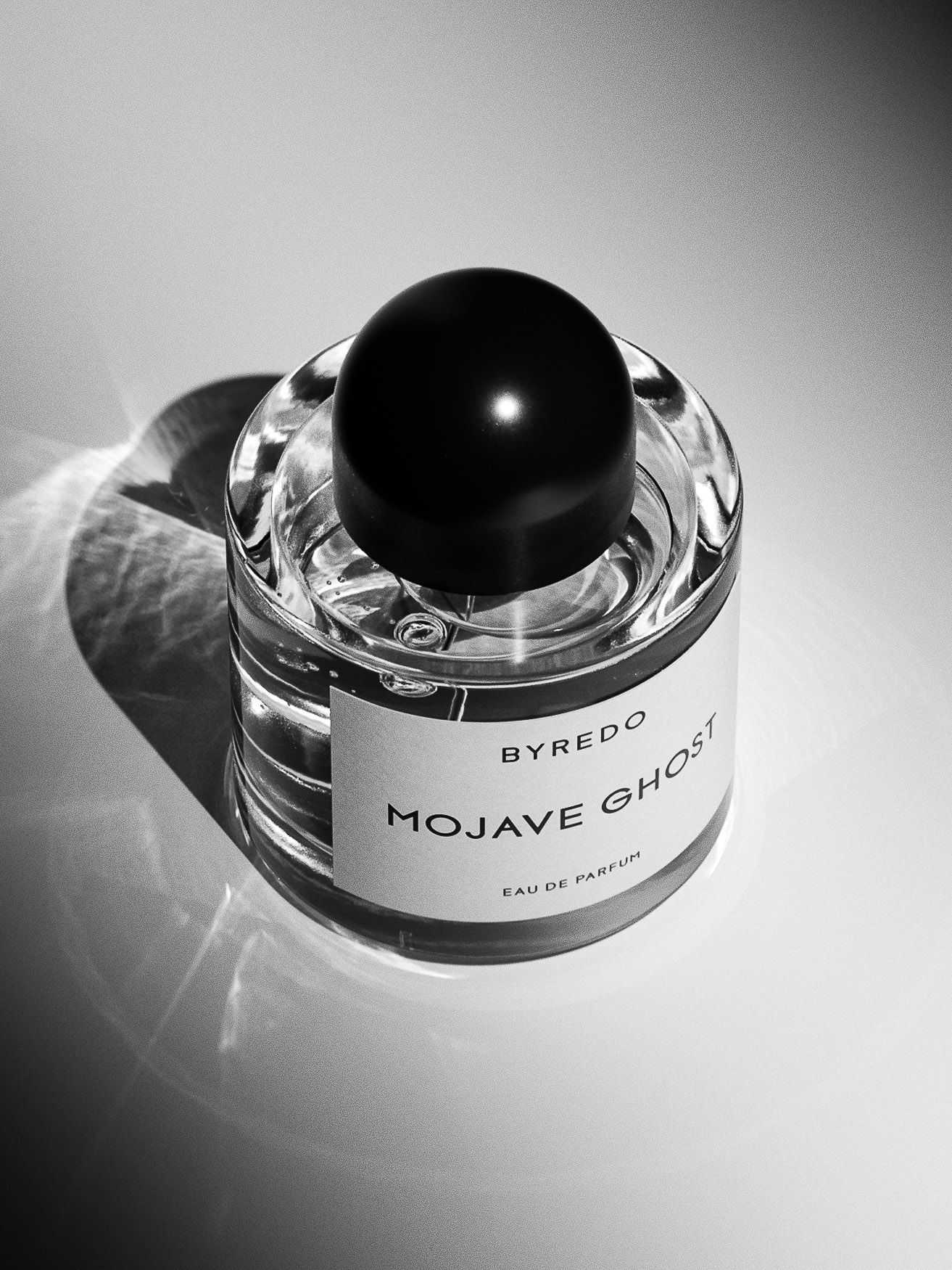 Black and white product shot of a clear glass bottle of Mojave Ghost Eau de Parfum by Byredo, with a round black cap and white label.