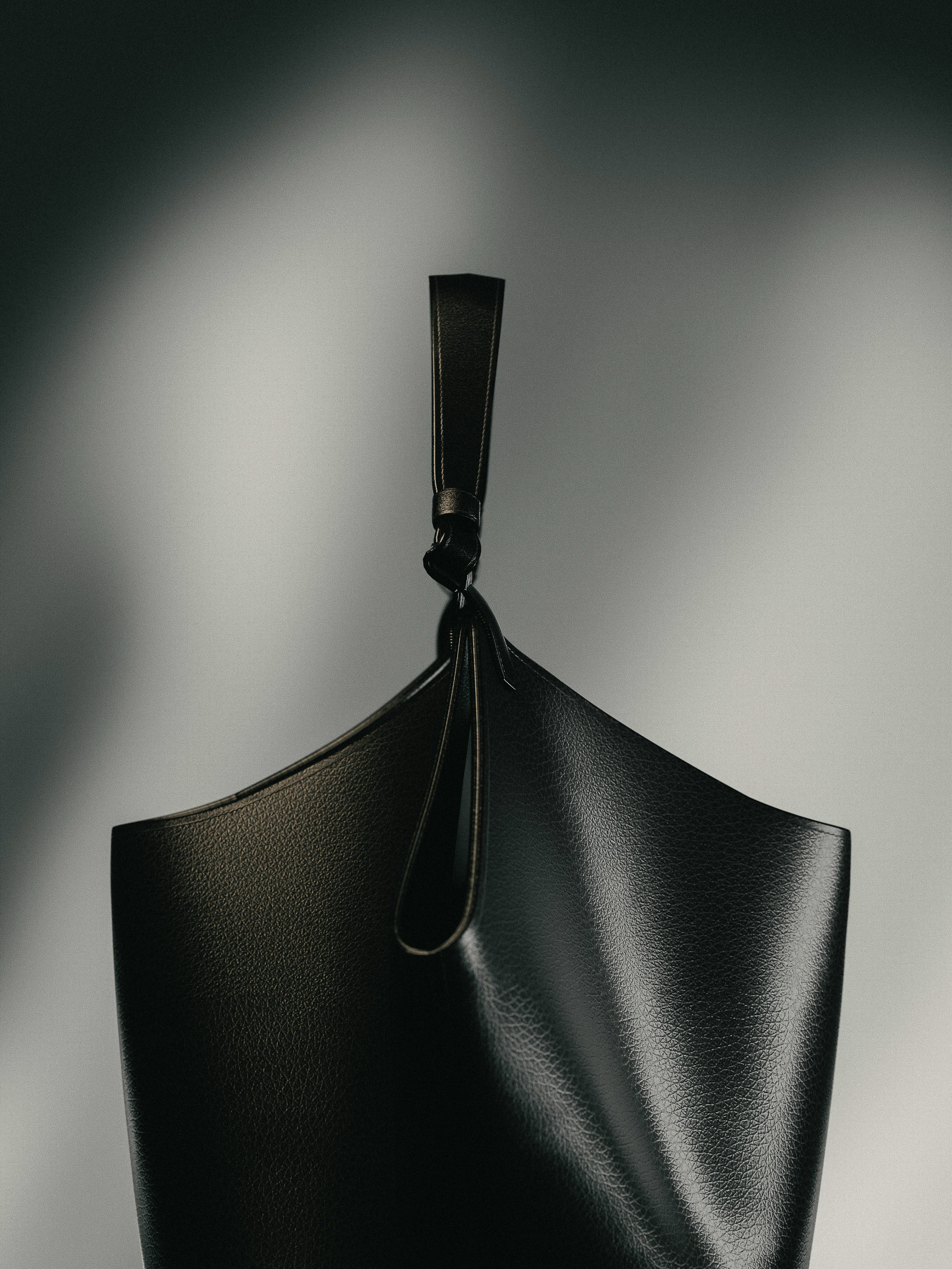 Stylish black leather bucket bag with unique knot detail, set against a soft grey gradient backdrop in a studio setting. Minimalist fashion.