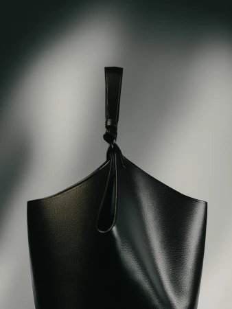 Stylish black leather bucket bag with unique knot detail, set against a soft grey gradient backdrop in a studio setting. Minimalist fashion.