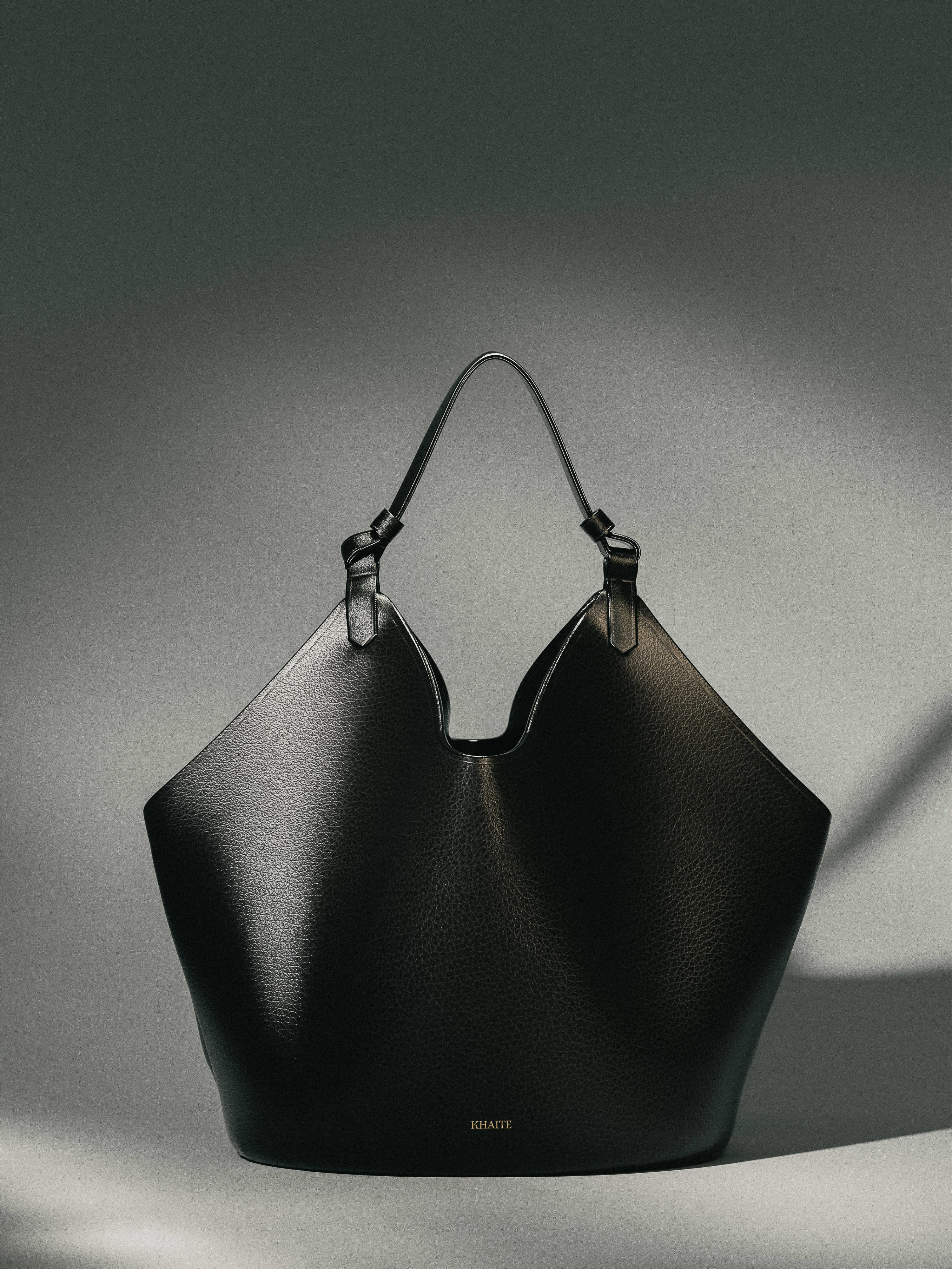 Black leather tote bag with top handles on a neutral background in a studio setting, product shot for fashion accessory, Khaite branding visible.