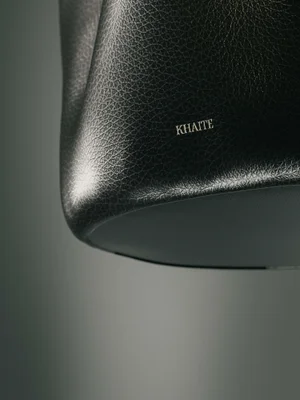 Close-up of a black textured leather Khaite handbag with metallic logo lettering against a gradient grey studio backdrop.