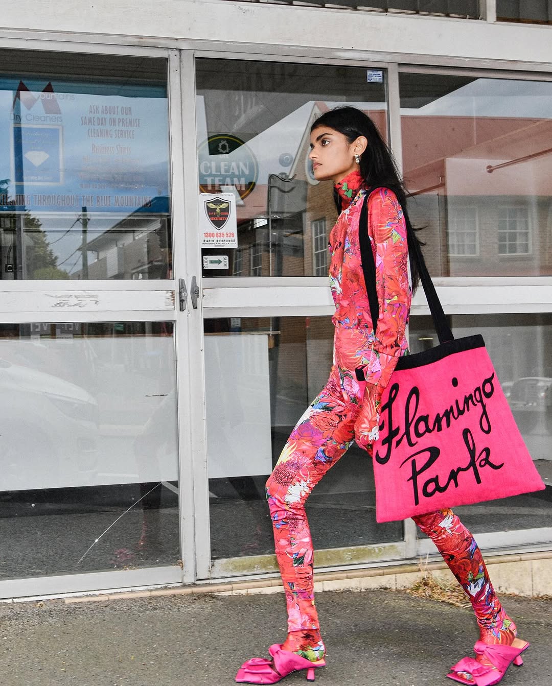 A woman in a vibrant pink patterned outfit and matching tote bag walks past an urban storefront.