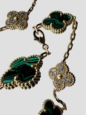 Close-up of a gold chain necklace featuring green malachite and diamond clover-shaped pendants on a neutral background, studio lighting.