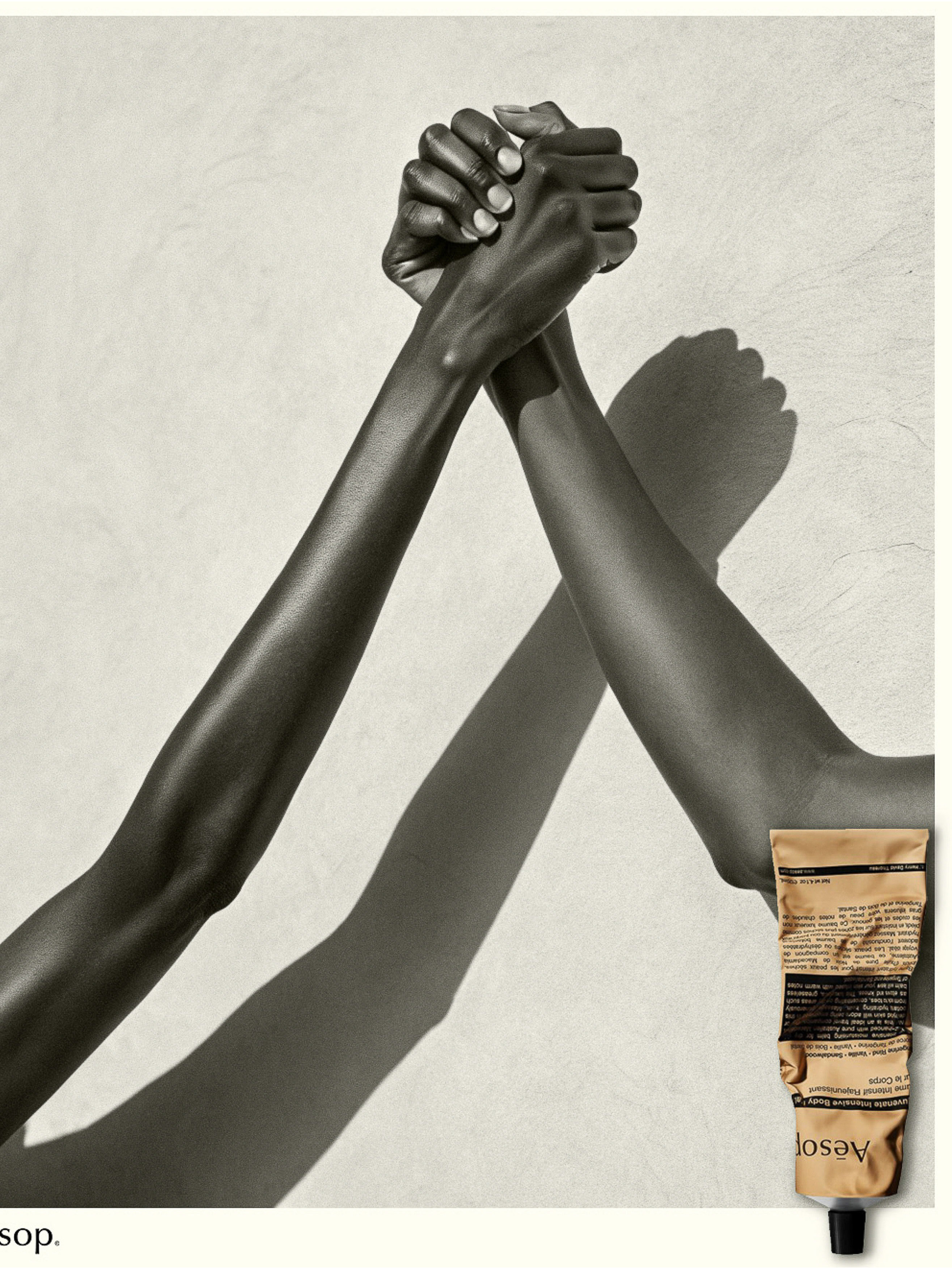 Close-up studio shot of two Black humans arm wrestling, showcasing a neutral-toned Aesop hand balm tube. The image has a clean, minimalist aesthetic.