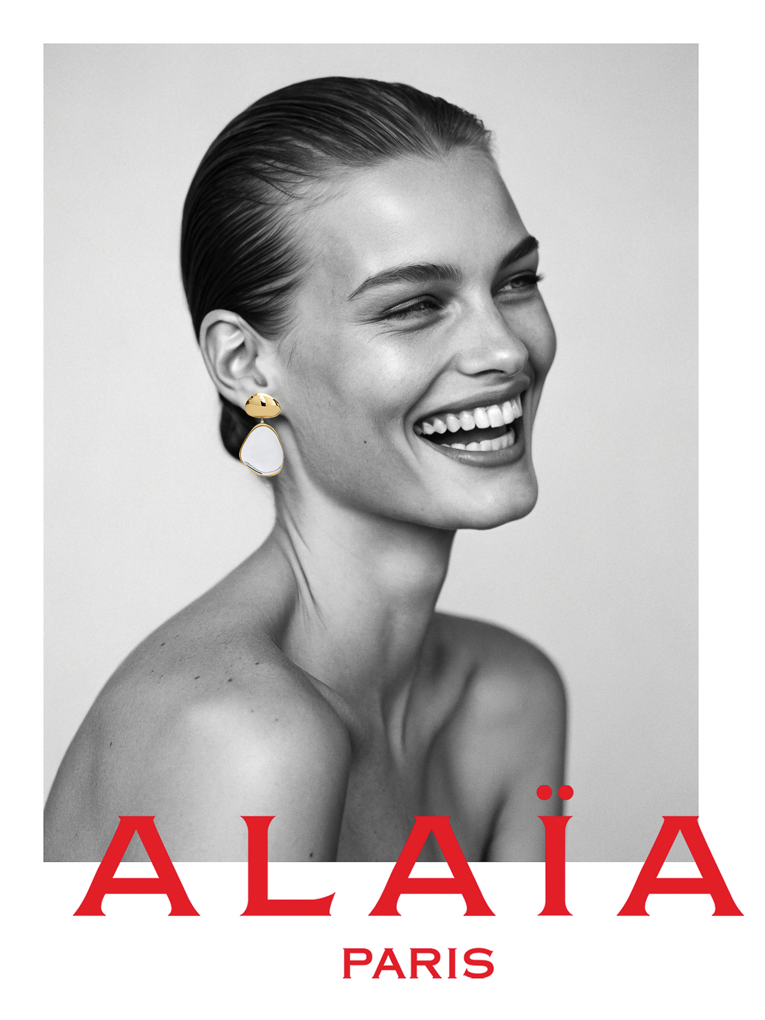 Close-up black and white portrait of smiling adult woman with gold and white earrings, clean face, and bare shoulders.
