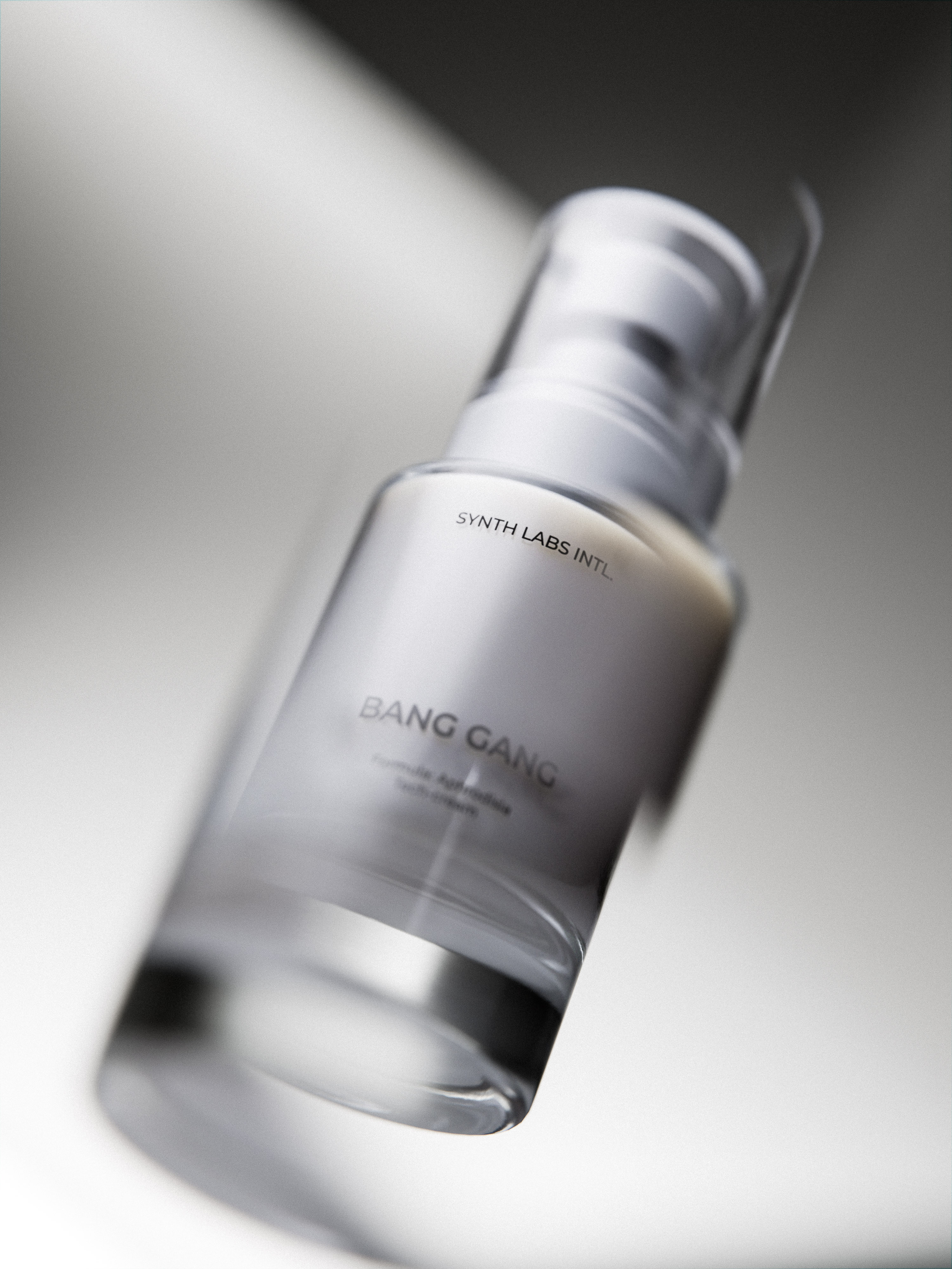 A clear glass bottle of Bang Gang Hydronic Aphrodisiac face cream stands against a blurred white background with reflected light.
