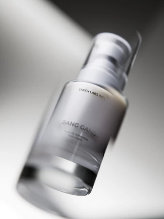 A clear glass bottle of Bang Gang Hydronic Aphrodisiac face cream stands against a blurred white background with reflected light.