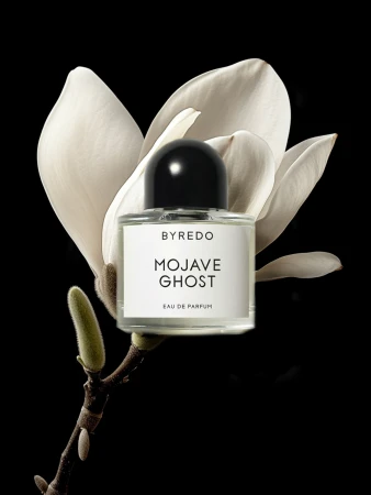 Byredo Mojave Ghost perfume bottle with black cap, white label, and magnolia flower on a black background in a studio shot.