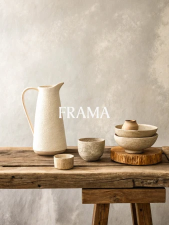 Still life composition featuring a ceramic pitcher, bowls, and cup arranged on a rustic wooden table against a neutral backdrop.
