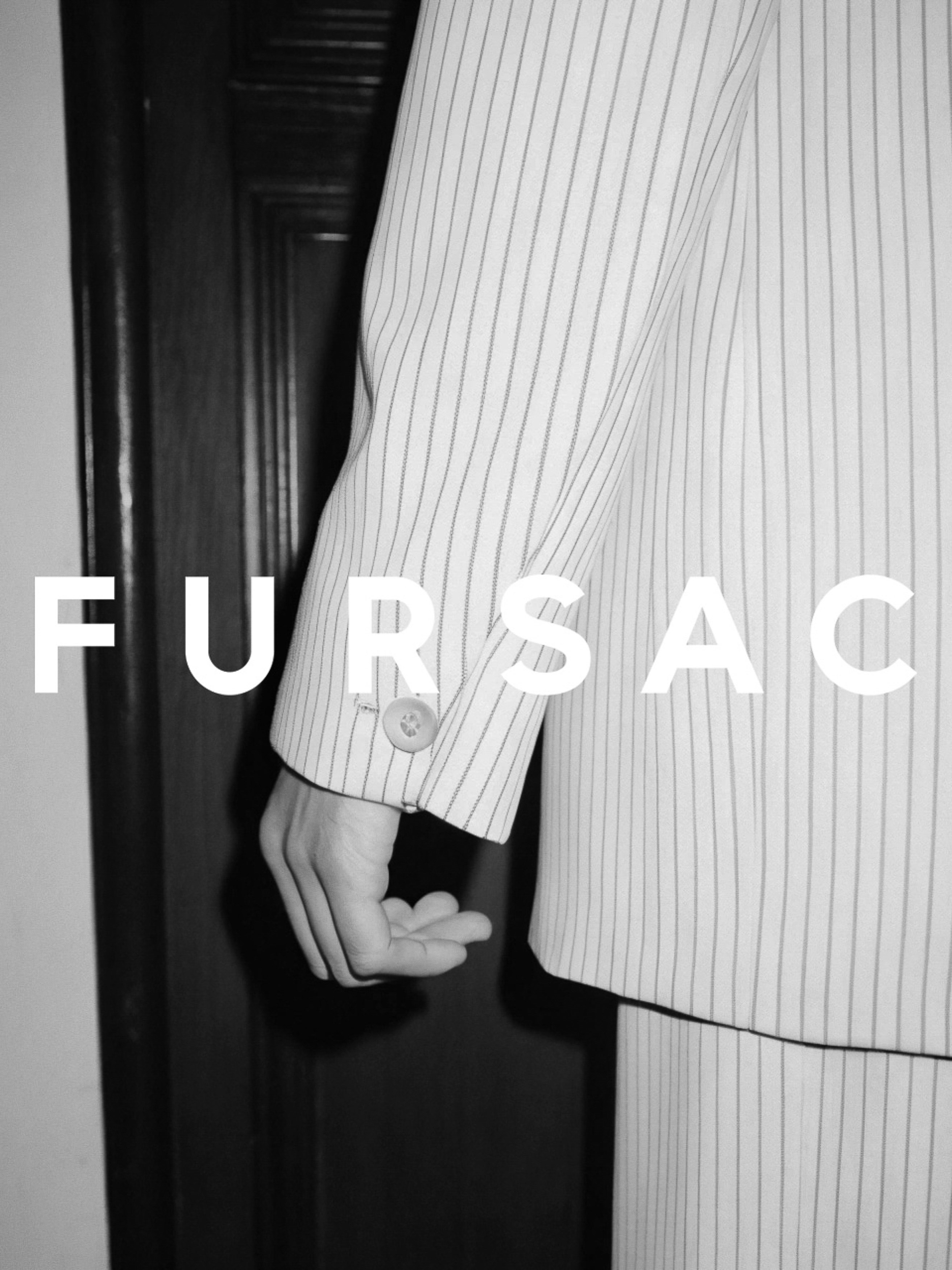 Close-up of a person's hand near a striped white suit jacket with a button in a monochrome studio setting with a partially visible black door.