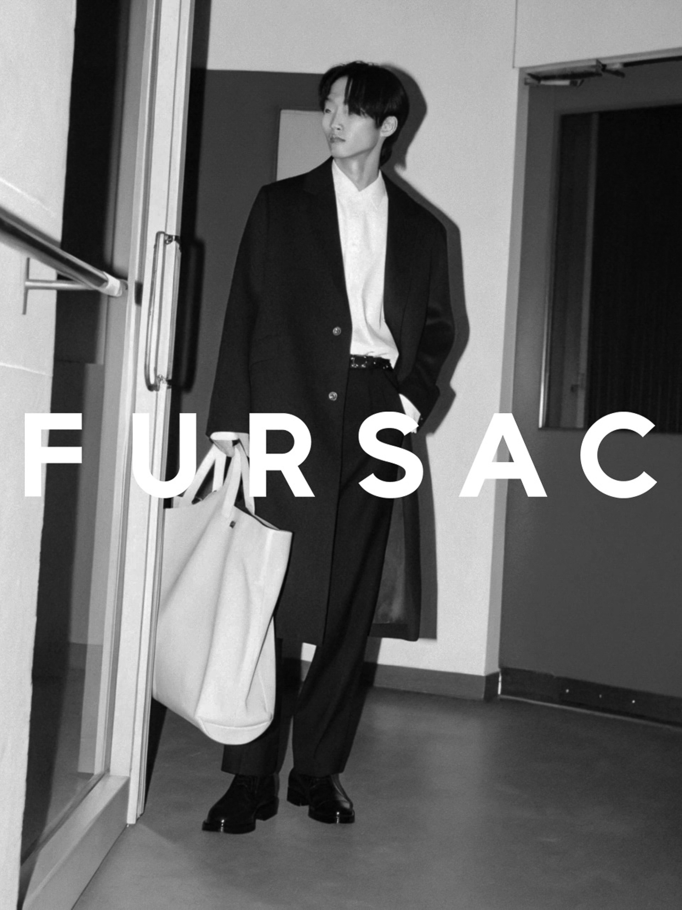 A black and white studio shot of a tall, slender male model wearing a long black coat, holding a tote bag, posing in a corridor.