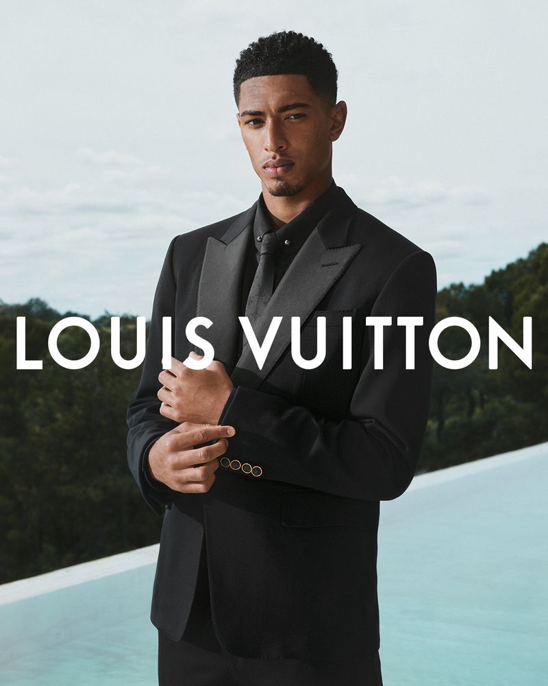 Man in black suit stands outdoors, Louis Vuitton logo visible. Sharp focus on subject, soft background, and modern fashion feel.