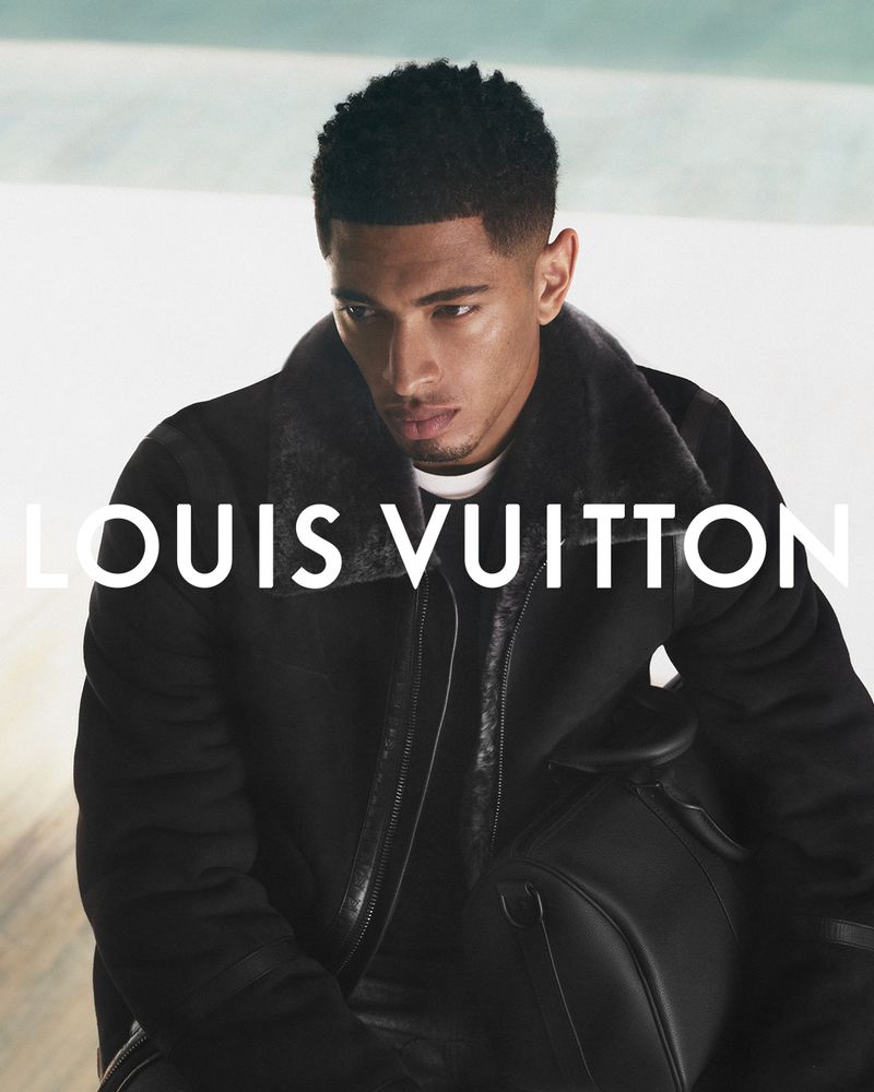 A young adult male model poses indoors wearing a black shearling coat with the Louis Vuitton logo visible. He has short dark hair.