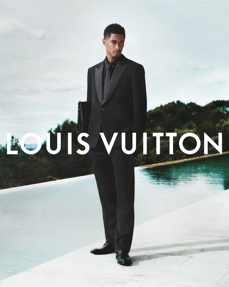 Fashion shot of a male model in a black suit and tie, standing near a pool with a forest background and Louis Vuitton logo.