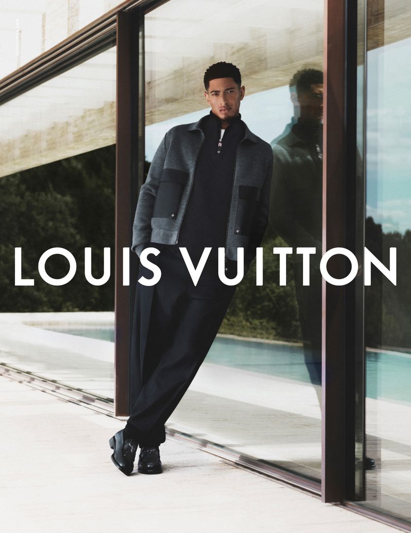 A stylish adult male model leans against a reflective glass surface in an outdoor fashion advertisement for Louis Vuitton, showcasing apparel.