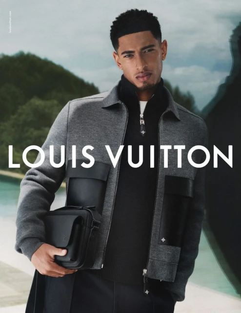 Man in grey jacket holding black bag against a blurred outdoor background, Louis Vuitton logo visible, stylish menswear fashion shoot.