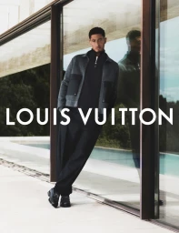 Jude Bellingham leaning against a modern glass building exterior wearing a grey jacket and black trousers for Louis Vuitton.