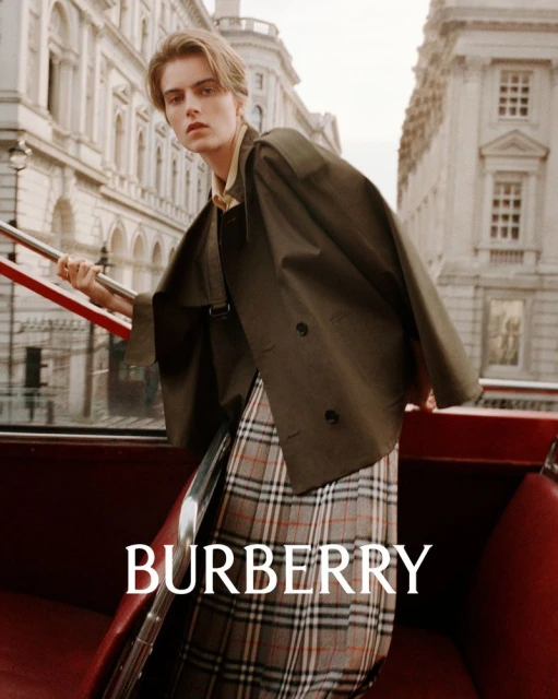 Male model wears olive green trench coat and plaid skirt on a red double-decker bus in front of a light stone urban building.