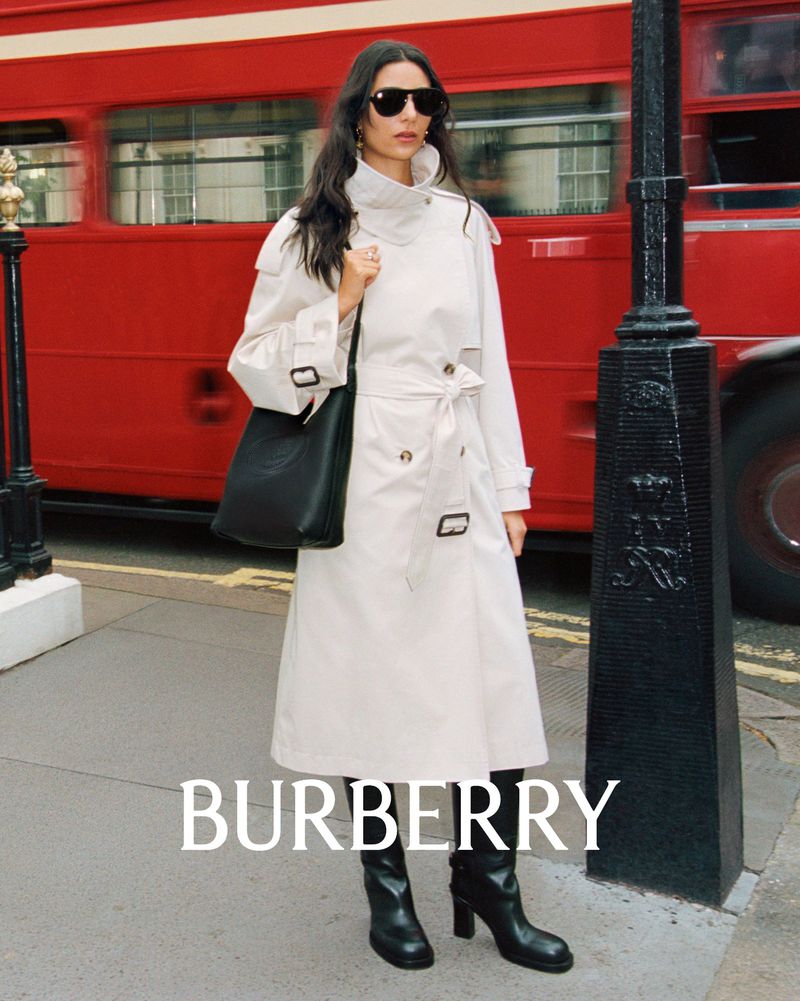 A woman models a beige trench coat and black boots on a city street with a red double-decker bus blurred in the background.