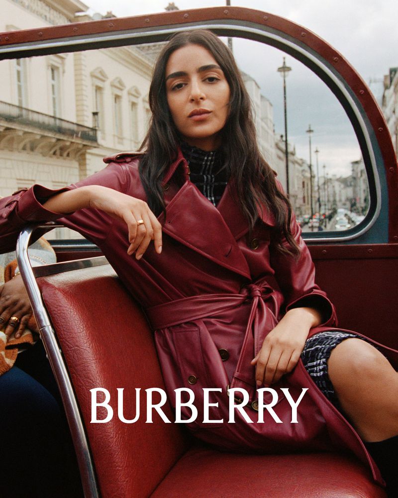 Brunette woman in a red trench coat sitting in a red bus seat, inside a double-decker bus in London with Burberry logo.