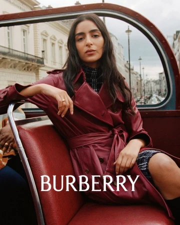 Brunette woman in a red trench coat sitting in a red bus seat, inside a double-decker bus in London with Burberry logo.