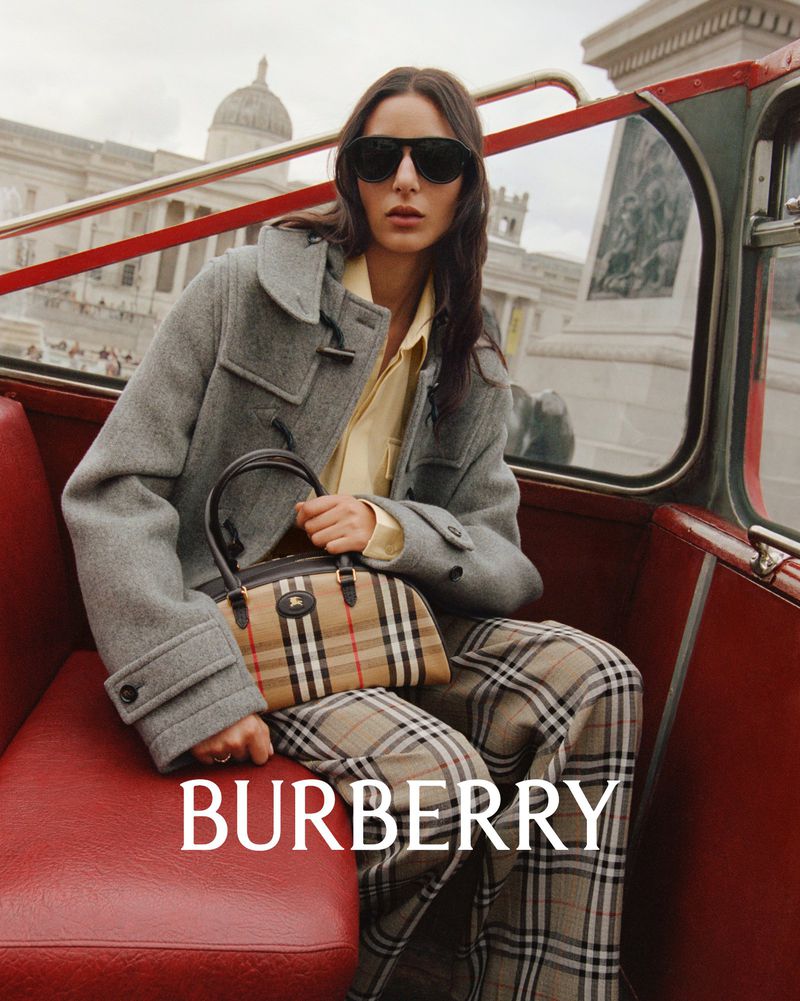 A young woman in a gray coat and plaid pants poses in a vintage red bus, holding a Burberry handbag, with London architecture visible.