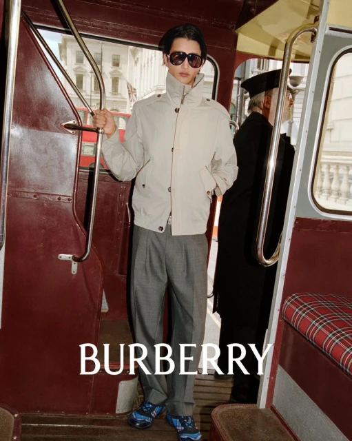 Man in neutral bomber jacket, grey trousers, and sunglasses stands on a vintage bus in a fashion editorial; Burberry logo at bottom.