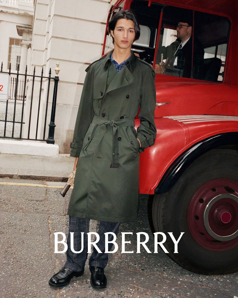 A young male model poses in a dark green trench coat against a red London bus and white building backdrop for a Burberry fashion shoot.