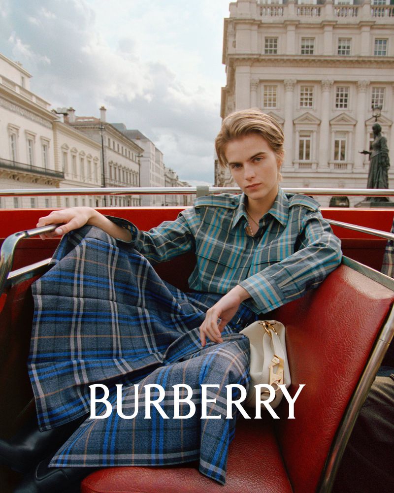 A woman wearing a plaid shirt and skirt sits on a red bus in front of a large building in a fashion advertisement, focusing on Burberry.