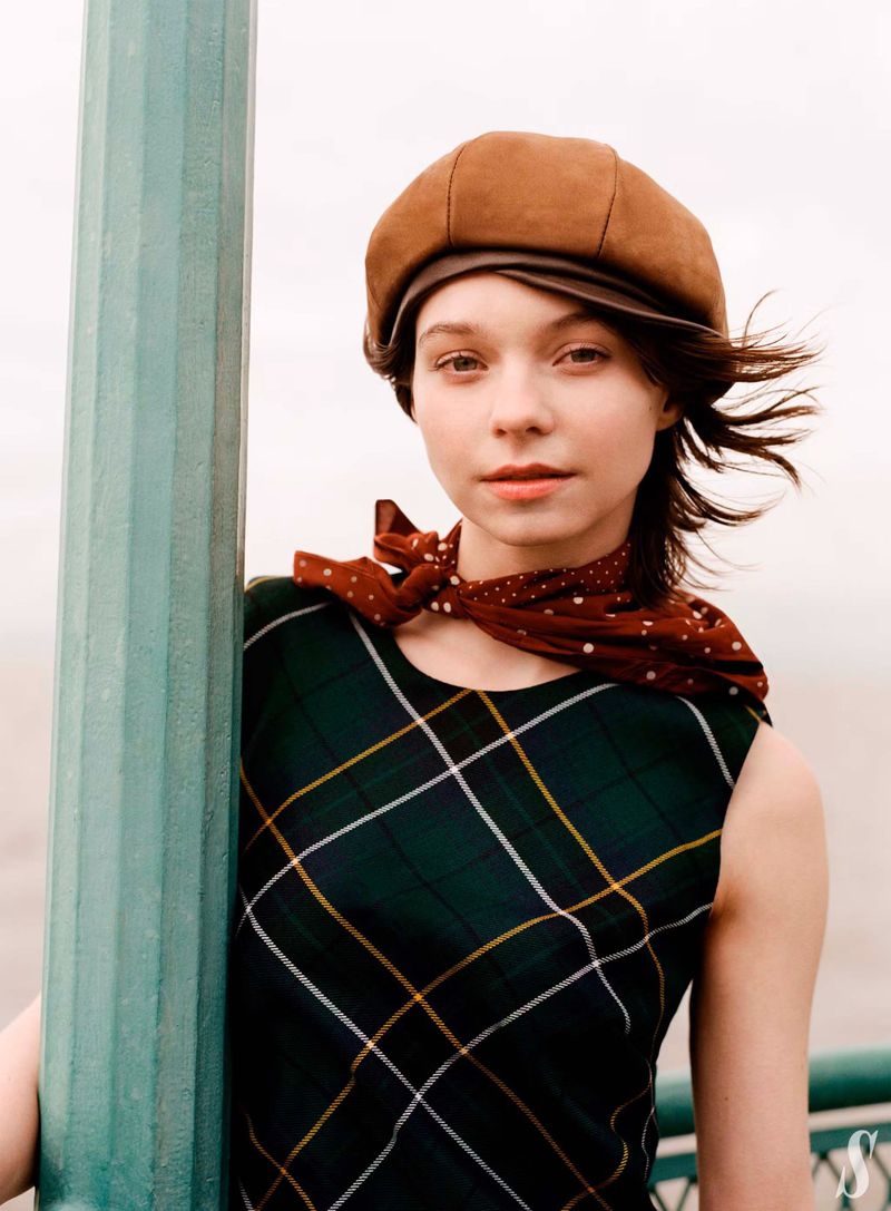 Portrait of a young woman wearing a green plaid dress, brown hat, and red scarf next to a light-blue pillar against a neutral backdrop.