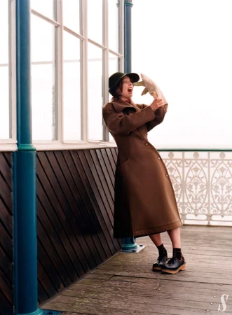 Woman in a brown coat and hat holds a fish while standing on a pier with railing and posts, exclaiming in a theatrical pose.