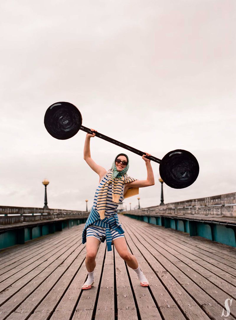 Woman in striped outfit and headscarf raises a barbell on a wooden pier with vintage lights under cloudy skies, fitness lifestyle.