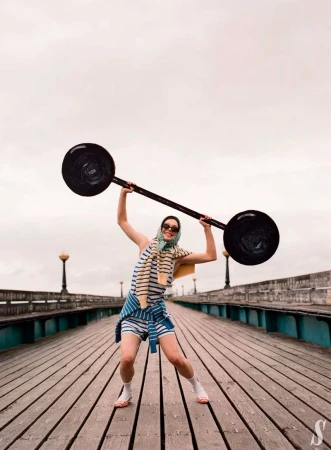 Woman in striped outfit and headscarf raises a barbell on a wooden pier with vintage lights under cloudy skies, fitness lifestyle.