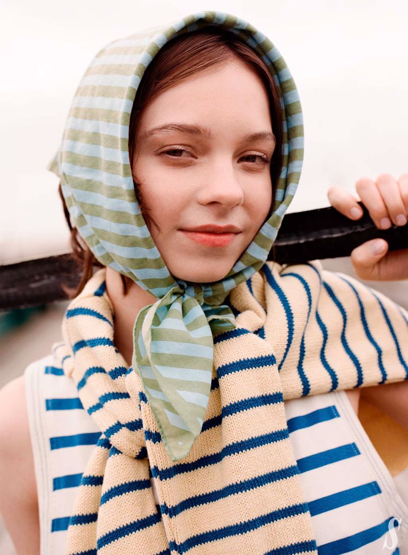 Portrait of a young woman wearing a striped headscarf and tank top, smiling softly in an outdoor setting with diffused lighting.