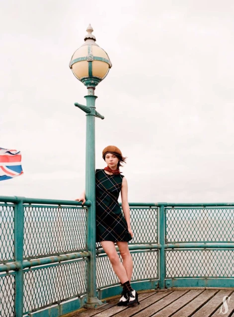 A young woman in a retro dress stands on a pier next to a vintage lamppost with a British flag waving in the background on an overcast day.