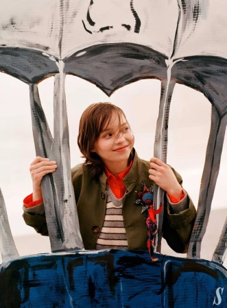 A smiling young woman in a green jacket and colorful shirt is posing behind an abstract, painted blue and white structure outdoors.