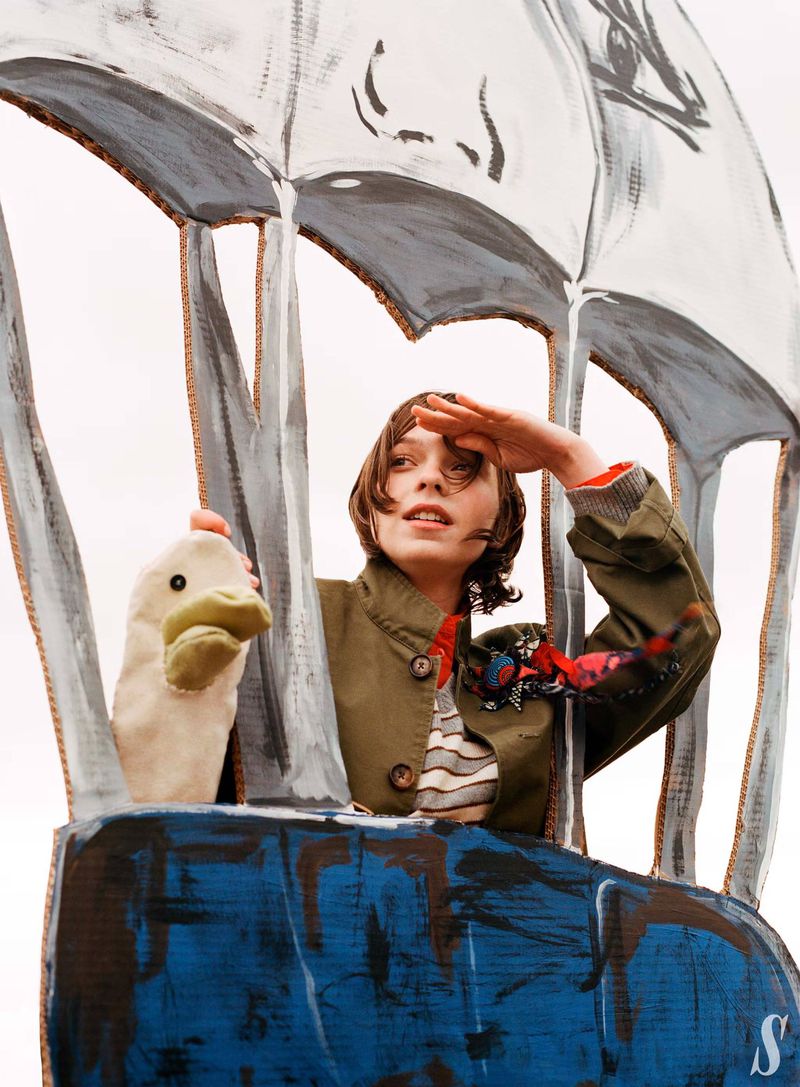 Adult female in a cardboard hot air balloon with a puppet, shielding her eyes with her hand, with a grey sky background.