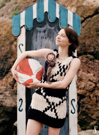 Adult female in a knit dress holds a red and white beach ball near a blue and white striped beach hut in an outdoor setting.