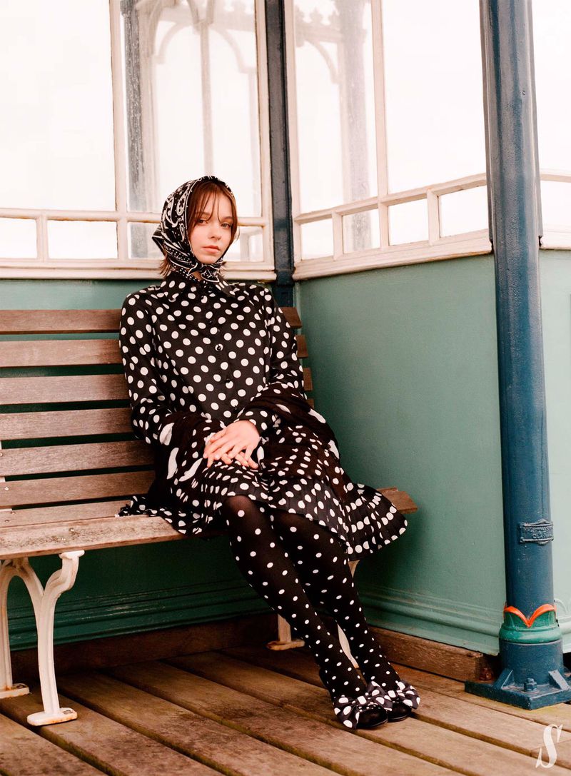 Woman in polka dot dress and headscarf sits on a weathered bench in a covered outdoor setting with light teal and cream accents.