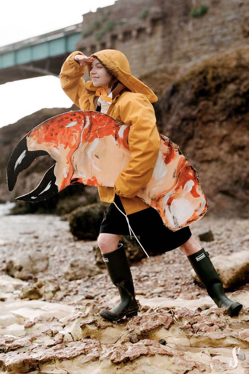 Woman in yellow raincoat, black skirt, and boots stands on a rocky beach, holding a large crab claw prop near a stone bridge.