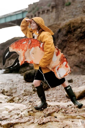 Woman in yellow raincoat, black skirt, and boots stands on a rocky beach, holding a large crab claw prop near a stone bridge.