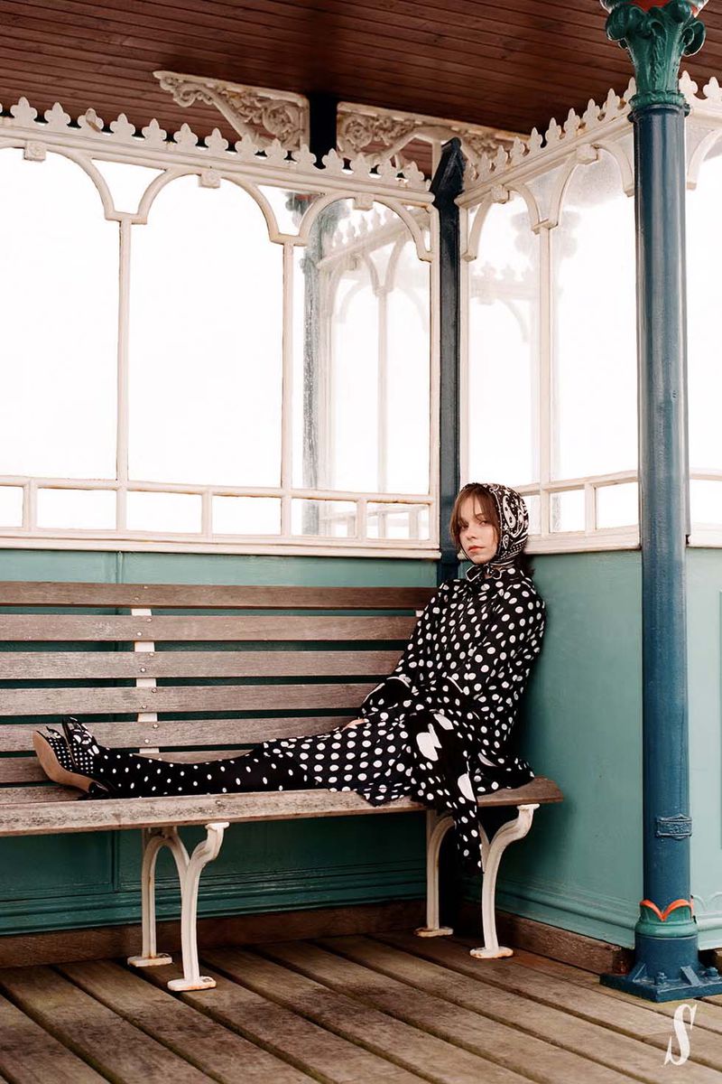 Young woman in a black and white polka dot outfit, including headscarf, poses on a bench under a vintage pavilion structure.