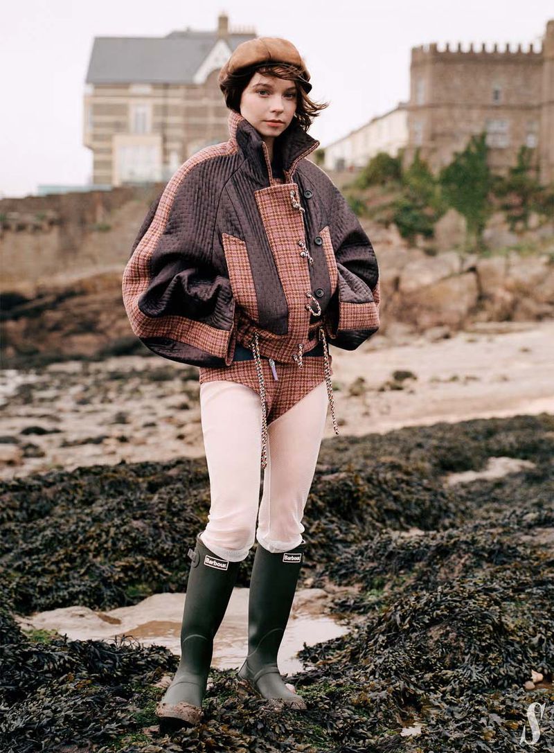 Fashionable young woman in jacket, beret, and rubber boots posing on rocky beach with buildings in background on an overcast day.