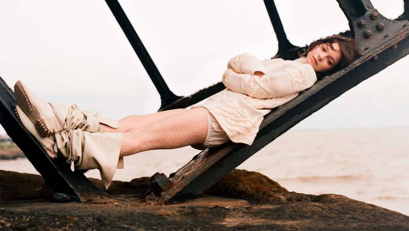 A young woman in a sweater and boots reclines on a metal structure outdoors with a neutral expression, set against a blurred background.