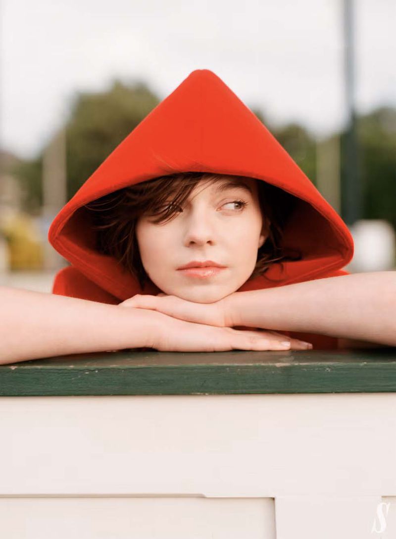 Portrait of a young woman wearing a bright red hooded cloak, resting her arms on a dark green ledge against an outdoor background.