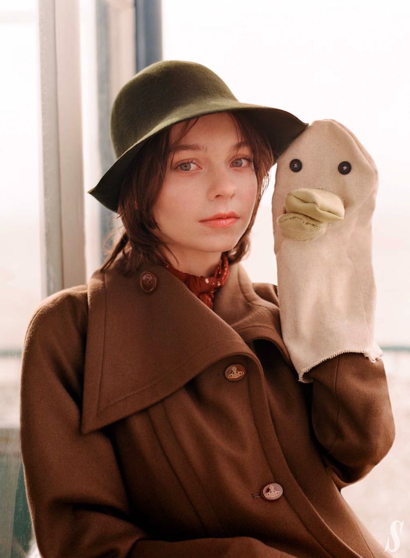 Young woman in a brown coat and olive hat stands next to a window, holding a duck hand puppet in a close-up studio-style shot.