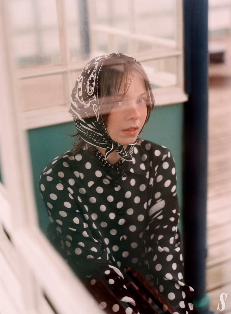 Young woman in a black-and-white polka dot shirt and paisley headscarf poses indoors near a window with diffused natural light.